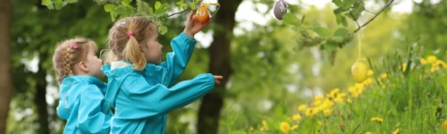 Kuvassa kaksi lasta poimii puuhub ripustettuja pääsiäismunia.